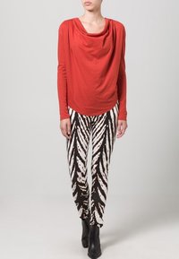 Czerwona bluzka z długim rękawem z materiału drapowanego w połączeniu z legginsami w czarno-białe paski zebra. Modelka nosi czarne botki za kostkę, stojąc na neutralnym tle.