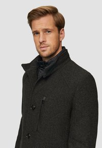 Zwarte textuurjasje met een hoge kraag, voorzien van een ritszak en twee knoopsluitingen. Het materiaal lijkt warm en zwaar te zijn.