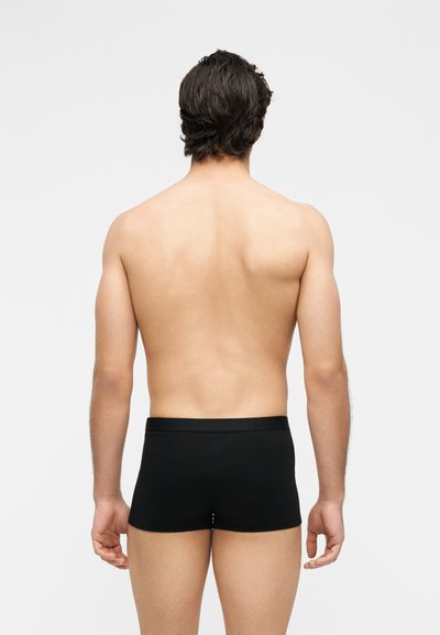 Άνδρας με σκούρα μαλλιά που φοράει μαύρα boxer briefs, στέκεται με την πλάτη στην κάμερα μπροστά σε απλό φωτεινό φόντο.
