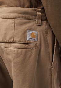 Κοντινό πλάνο μπεζ παντελονιού Carhartt που δείχνει την πίσω τσέπη με το λογότυπο και τη θηλιά ζώνης στη μέση.