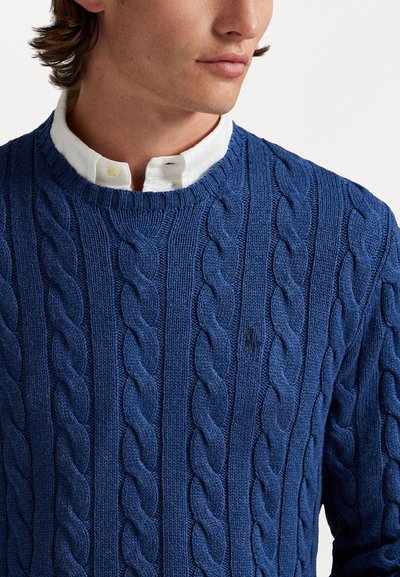 Μπλε πλεκτό πουλόβερ με πλέξη καλωδίων και στρογγυλή λαιμόκοψη, συνδυασμένο με λευκό πουκάμισο από κάτω. Μικρή λεπτομέρεια λογότυπου στο στήθ�ος.