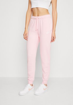 Moteris vilkinti šviesiai rožinius Nike treninginius kelnes su raišteliu ir baltus Nike sportbačius prieš neutralų foną.