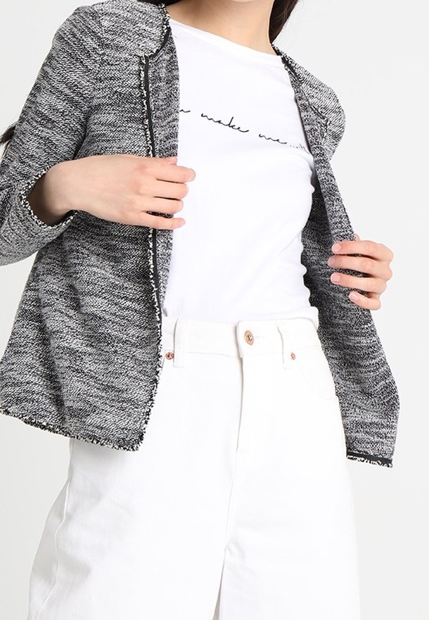 Femme portant une veste grise texturée ouverte sur un t-shirt blanc graphique et un pantalon blanc taille haute avec des boutons.