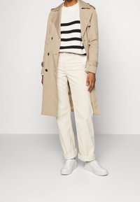 Beige trenchcoat päällä mustavalkoista raidoitettua neuletta, yhdistettynä käännettyihin kermaisiin housuihin ja valkoisiin lenkkareihin. Sileät kangasrakenteet.