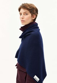 Persoon met kort haar die een donkerblauwe sjaal over een bordeauxrode jas draagt, zijwaarts gekeerd met een neutrale uitdrukking op een effen achtergrond.