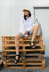 Sandalias negras con correas ajustables, llevadas con pantalones cortos grises y una camisa blanca, sentadas sobre palets de madera, acompañadas de un bolso de estampado de leopardo.