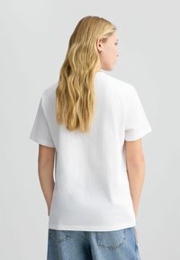 T-shirt en coton blanc avec une coupe décontractée, des manches courtes et un col rond. Texture lisse sans motifs ni accents visibles.