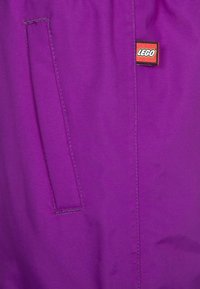 Fioletowa tkanina ma naszytą boczną kieszeń oraz metkę z logo LEGO. Tekstura wydaje się gładka, a kolor jest intensywny.