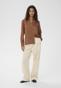 Braunes, gepunktetes Hemd mit spitzem Kragen, Knopfleiste vorne und langen Ärmeln, kombiniert mit cremefarbenen, weit geschnittenen Hosen und schwarzen Schuhen.