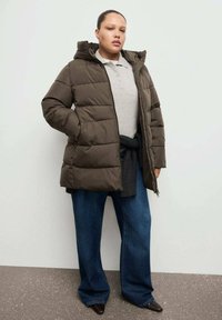 Ciemnozielona pikowana kurtka z kapturem, zapinana na zamek z przodu, noszona na szarej koszulce polo. Do tego szerokie niebieskie dżinsy i spiczaste czarne buty.