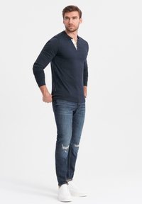 Hombre con una camiseta henley marina, jeans azules desgastados y zapatillas blancas, de pie con las manos cerca de los bolsillos, mirando de lado sobre un fondo blanco.