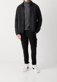 Černá bunda na zapínání přes knoflíky přes šedý pletený rolák, doplněná černými kalhotami s kapsami a bílými vysokými teniskami s černými detaily.