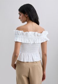 Kvinna med långt mörkt hår som bär en vit off-shoulder topp med volanger och beige byxor, vänd åt vänster mot en neutral bakgrund.