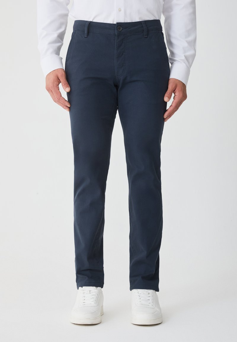 Námornícke modré chino nohavice so slim fit, vyrobené z bavlny, s bočnými vreckami, predným zapínaním na gombík a v kombinácii s bielymi teniskami.