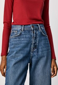 Person med en ribbad, långärmad röd topp instoppad i högmidjade blå denimjeans med synliga knappar och framfickor.