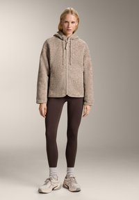 Beżowa polarowa kurtka zapinana na zamek z kapturem, charakteryzująca się miękką fakturą i przednimi kieszeniami. Noszona z brązowymi legginsami i jasnymi trampkami.