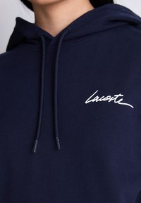Asmuo, vilkintis tamsiai mėlyną gobtuvą su baltu "Lacoste" logotipu kairėje krūtinės pusėje ir tamsiai mėlynomis raištelėmis, kabančiomis žemyn.
