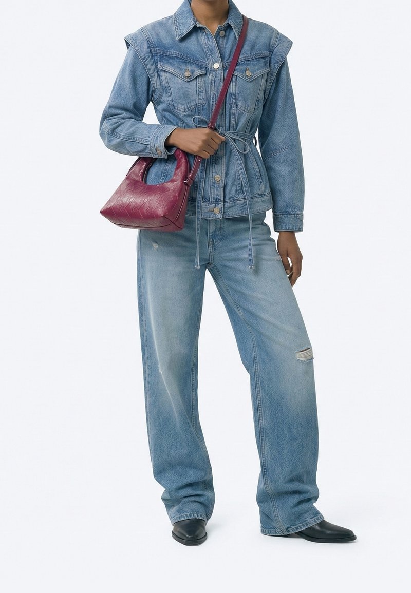 Personne portant une veste en jean bleue et un jean jambes larges tenant un petit sac à main en cuir rouge avec une bandoulière.