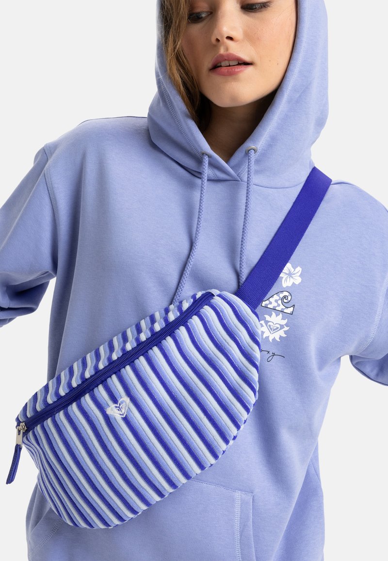 Blue and white striped fanny pack with textured fabric, contrasting blue strap, and zipper closure. Worn over a light purple hoodie.