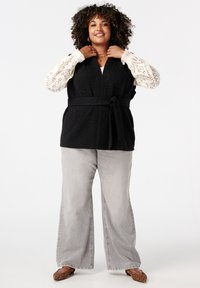 Zwarte, gestructureerde mouwloze vest met een strikceintuur, gecombineerd met een witte kant lange mouw top en grijze wide-leg jeans. Bruine schoenen met luipaardprint.