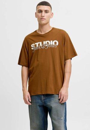 T-shirt en coton marron avec une coupe ample. Présente un texte blanc "STUDIO Jack & Jones" imprimé sur le devant. Design décontracté avec des manches courtes.