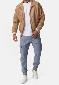 Man som bär en beige bomberjacka, vit T-shirt, ljusblå cargobyxor och vita sneakers, som står mot en neutral bakgrund.