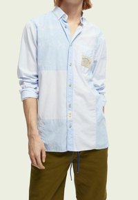 Camicia azzurro chiaro a maniche lunghe con motivi misti, caratterizzata da righe e pannelli solidi. Include una tasca sul petto con un'etichetta.