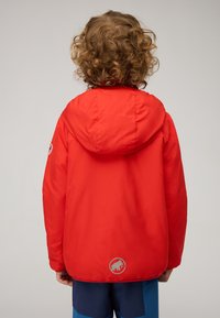 Giacca rossa con cappuccio dalla finitura liscia e texture. Presenta una toppa con il logo sulla schiena e cuciture semplici, abbinata a pantaloni corti blu.