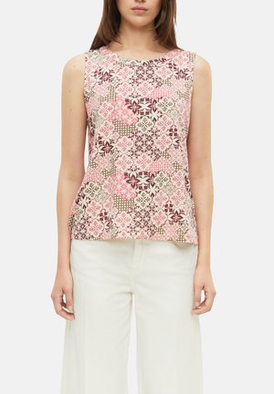 Femme portant un haut sans manches avec des motifs floraux et géométriques roses, beiges et marron, associé à un pantalon blanc taille haute.