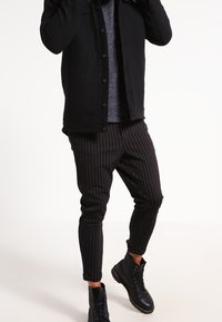 Chaqueta negra de botones sobre una camisa gris, combinada con pantalones negros a rayas y botas negras con cordones. Tela suave y diseño casual.