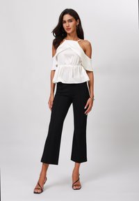 Haut blanc décolleté avec une texture plissée et une taille peplum, associé à un pantalon noir évasé. Le mannequin porte des talons noirs à lanières.