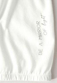 Weißes Baumwollkleidungsstück mit elastischen Bündchen, bedruckt mit dem Text "SEI EIN SPIEGEL DES Lichts" in metallischem Grau. Glatte Textur, lässiges Design.