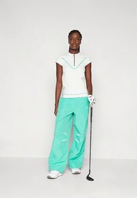 Γυναίκα φορώντας λευκή μπλούζα με κοντά μανίκια και Τουρκουάζ παντελόνι, κρατώντας ένα κλαμπ γκολφ με ένα λευκό γάντι στο δεξί της χέρι, στέκεται μπροστά σε απλό φόντο.