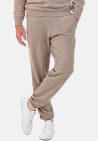 Joggers en maille beige en tissu doux, avec une ceinture élastique et des poches latérales, assortis à des baskets blanches sur un fond uni.
