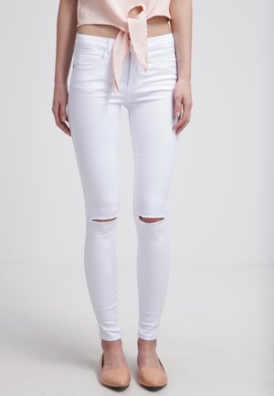 Vrouw draagt witte skinny jeans met scheuren bij de knieën, een roze top met knoop, en nude platte schoenen, staand tegen een effen achtergrond.