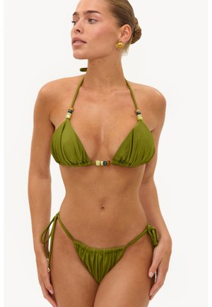 Femme portant un bikini vert avec des détails en perles, noué au niveau du cou et des hanches, posant devant un fond clair uni.