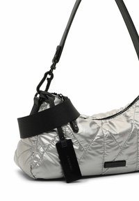 Bolso de mano acolchado en plata con acabado brillante, que presenta una correa y acentos negros, cierre de cremallera y una etiqueta rectangular de la marca.