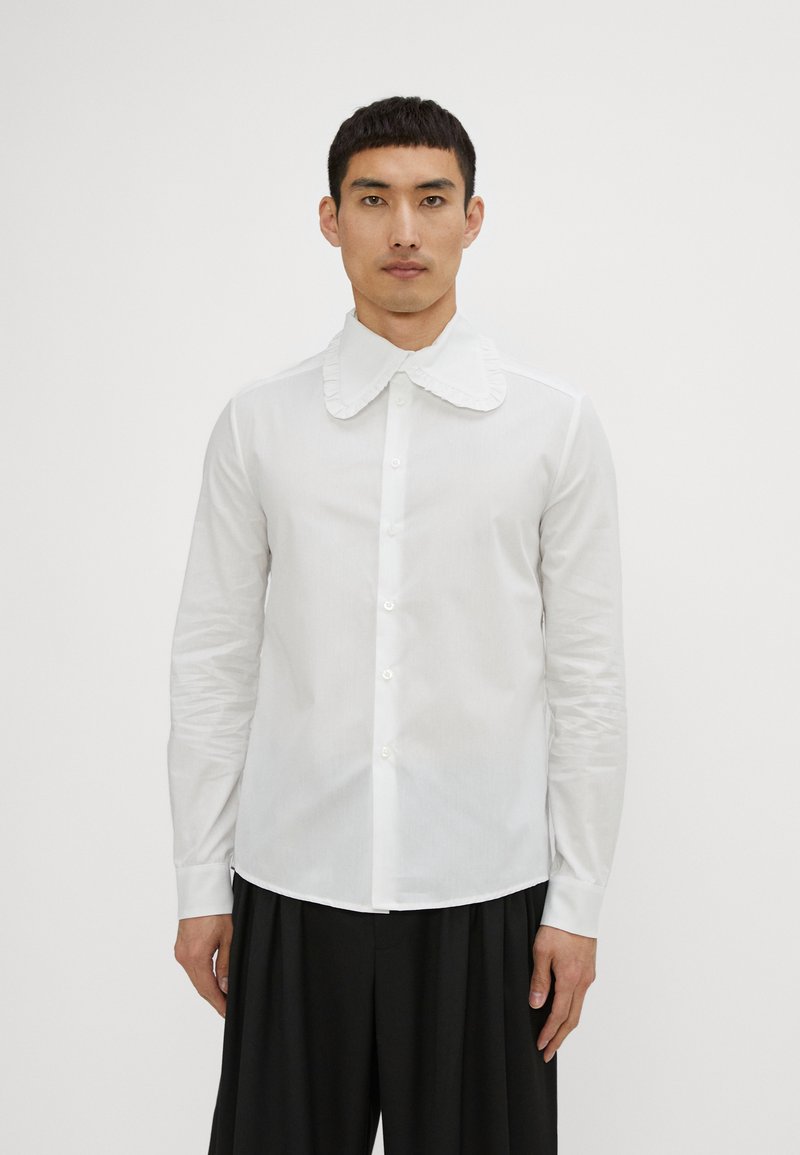 White button-up shirt with a ruffled collar, long sleeves, and a smooth texture, paired with black pleated pants.