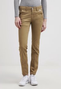 Beige bomullschinos med slim fit, med framfickor, knäppning med knapp och en liten metallisk logotyp på midjebandet. Bärs med vita sneakers.