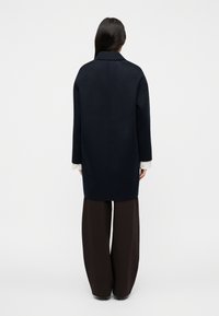 Ναυτικό μάλλινο παλτό με ίσια γραμμή, μεγάλο γιακά και μακριά μανίκια, που φοριέται πάνω από καφέ φαρδιές παντελόνες, με μινιμαλιστικό σχεδιασμό.