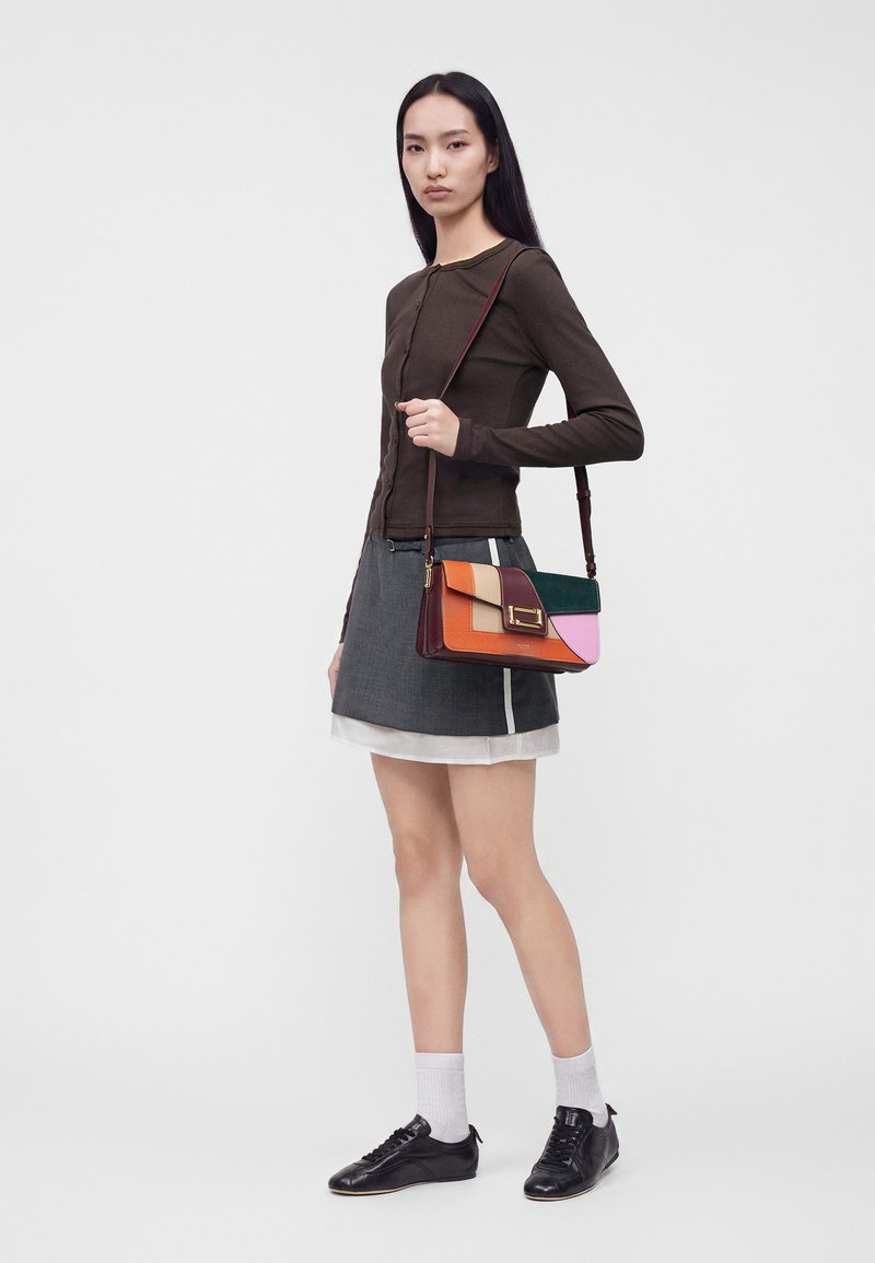 Femme portant un haut marron foncé à manches longues, une jupe grise, des chaussettes blanches, des chaussures noires, tenant un sac à bandoulière multicolore sur un fond uni.