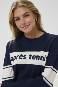 Námořnický modrý svetr s bílými vodorovnými pruhy; na hrudi se nachází nápis "après tennis" v tučném písmu. Měkká pletená textura.