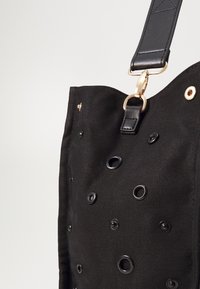 Sac fourre-tout noir en tissu texturé avec de multiples œillets, une fermeture en métal doré et une bandoulière en cuir noir.