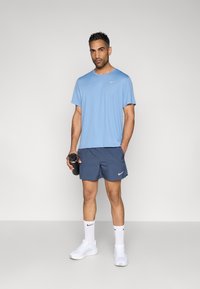 Világoskék Nike póló, tengerész kék rövidnadrág, fehér Nike zokni és fehér sportcipő. Egy fekete vízpalackot tart, semleges háttér előtt áll.