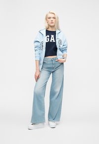 Junge Frau in hellblauer Kapuzenjacke, dunkelblauem GAP-Bauchfreitop, weit geschnittenen hellen Jeans und weißen Plateausneakers steht vor einem einfarbigen Hintergrund.