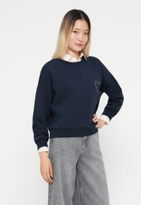 Felpa blu navy in cotone con colletto tondo, spalle cadenti e un piccolo design a cuore sul lato sinistro del petto. Abbinata a pantaloni a gamba larga grigi.