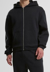 Uomo che indossa un felpa nera con zip e tasche frontali, abbinata a pantaloni neri, su uno sfondo neutro.