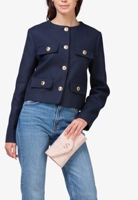 Marineblauwe cropped jas met gouden knopen en opgestikte zakken, gecombineerd met blauwe jeans. Houdt een lichtroze clutch met een subtiel logo vast.