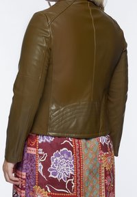 Dark olive green leather jacket with fitted design, quilted back panel, and smooth texture. Worn over a colorful patterned skirt.