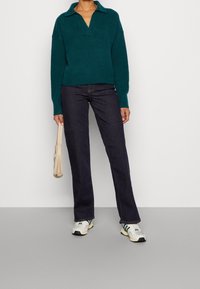 Osoba v tmavě modrých džínech, tyrkysovém svetru s dlouhým rukávem a bílých teniskách, držící béžovou kabelku na jednobarevném světlém pozadí.